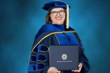 Woman in university regalia holding a diploma in front of a blue background.